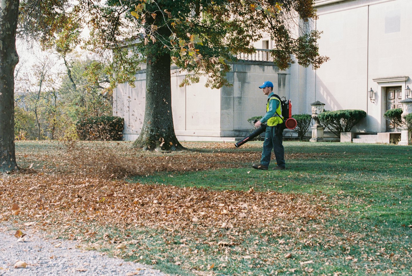 Groundskeeper clearing autumn leaves from a lawn near a stone building.